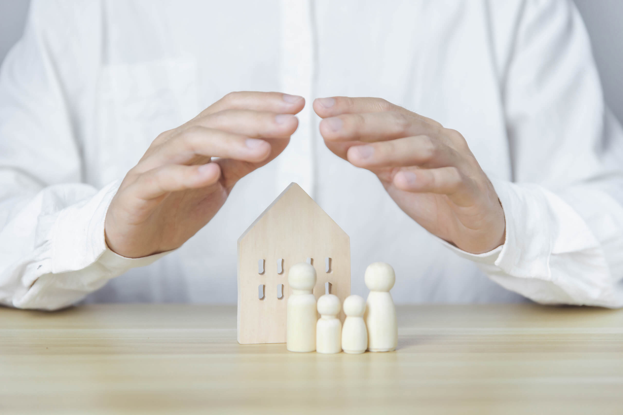 Hände schützen ein Holzhaus und Familienfiguren auf einem Tisch.