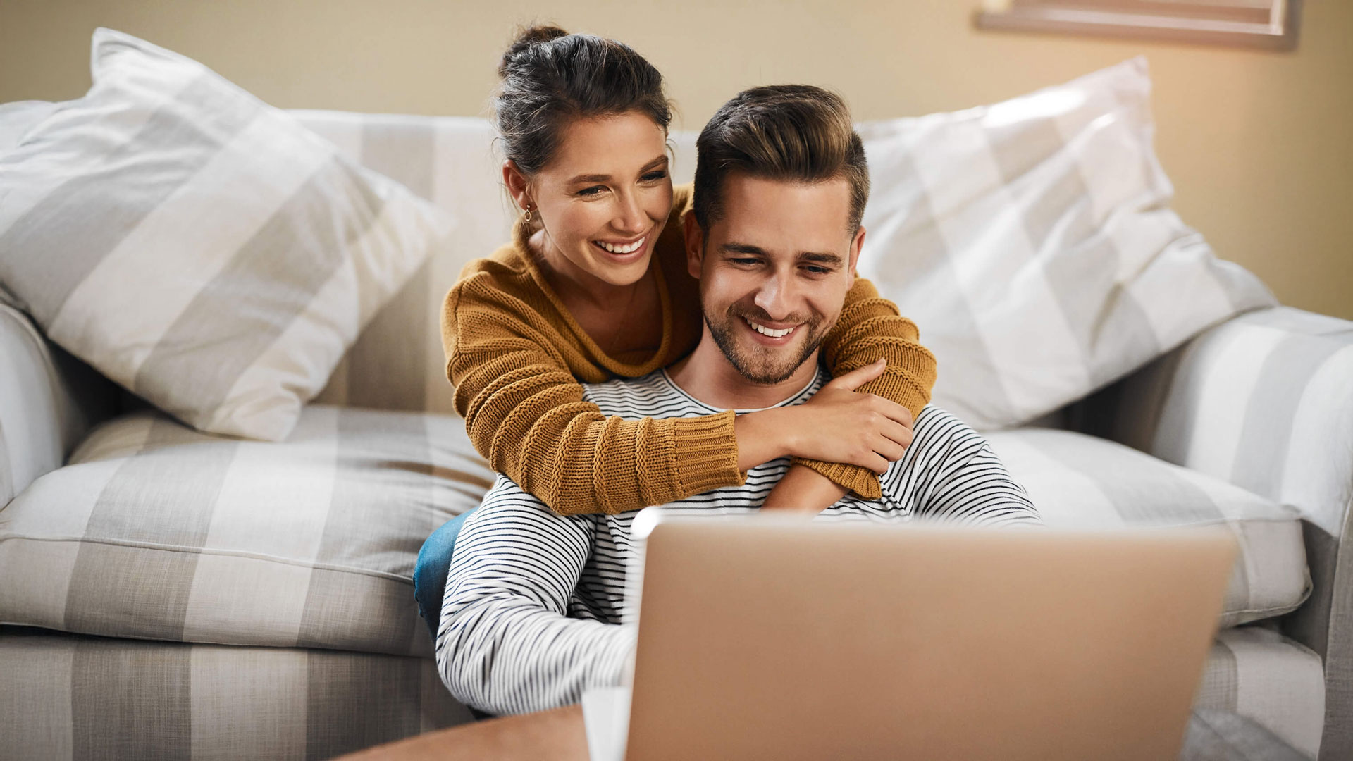 Paar sitzt am Boden, umarmt sich und schaut auf einen Laptop.