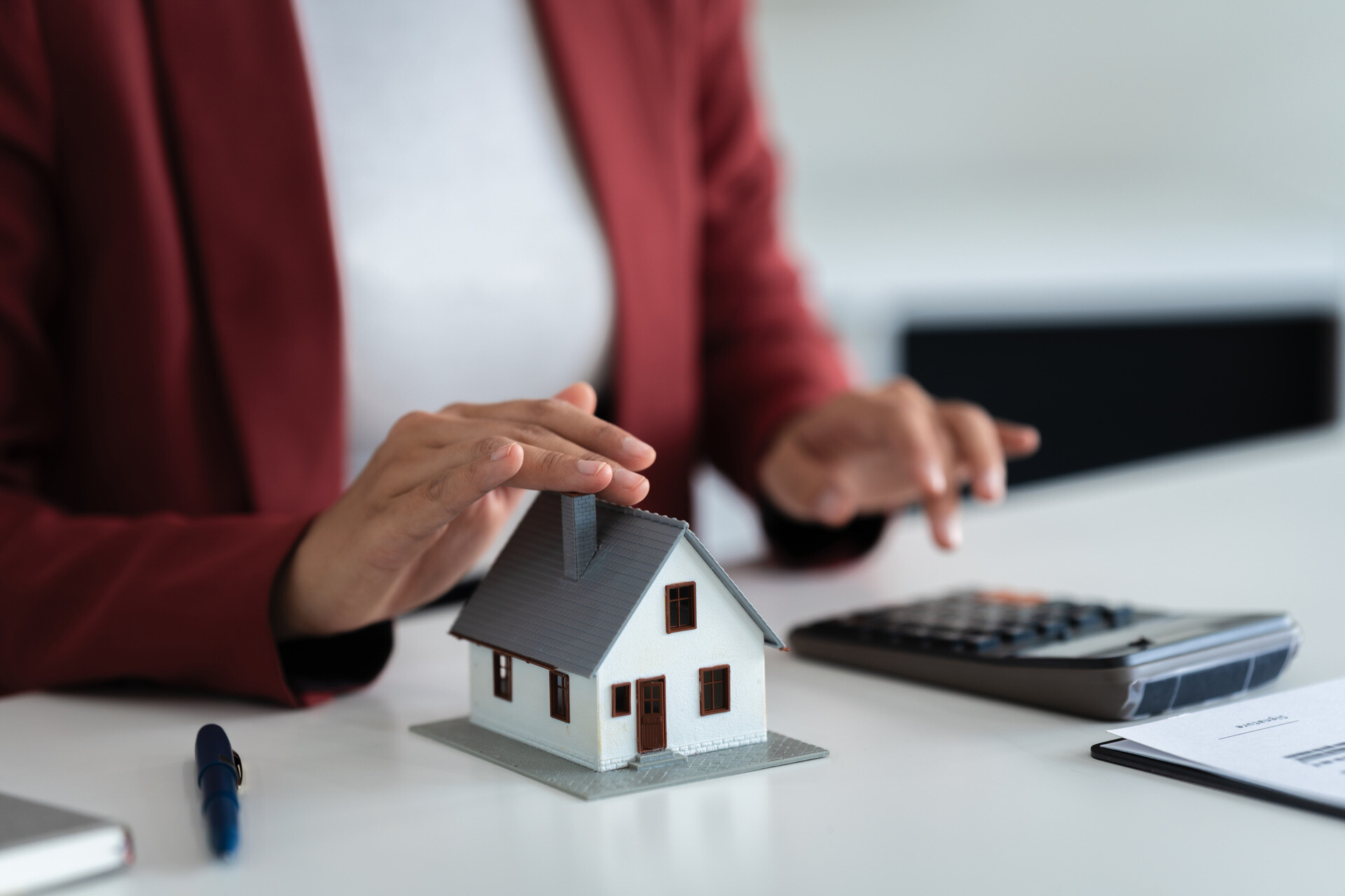 Frau tippt am Taschenrechner und hält ihre andere Hand über einem Hausmodell