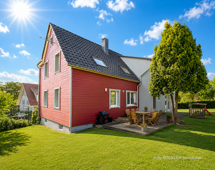 Energetisch saniertes Fachwerkhaus mit roter Holzfassade an einem sonnigen Sommertag. Die Fassade wird von hellem Sonnenlicht angestrahlt, der gepflegte Garten mit frisch gemähtem Rasen und die aufgeräumte Terrasse unterstreichen den Wohnkomfort. Das Bild verdeutlicht, wie eine energetische Sanierung auch bei hohen Außentemperaturen für ein angenehmes Raumklima sorgen kann.