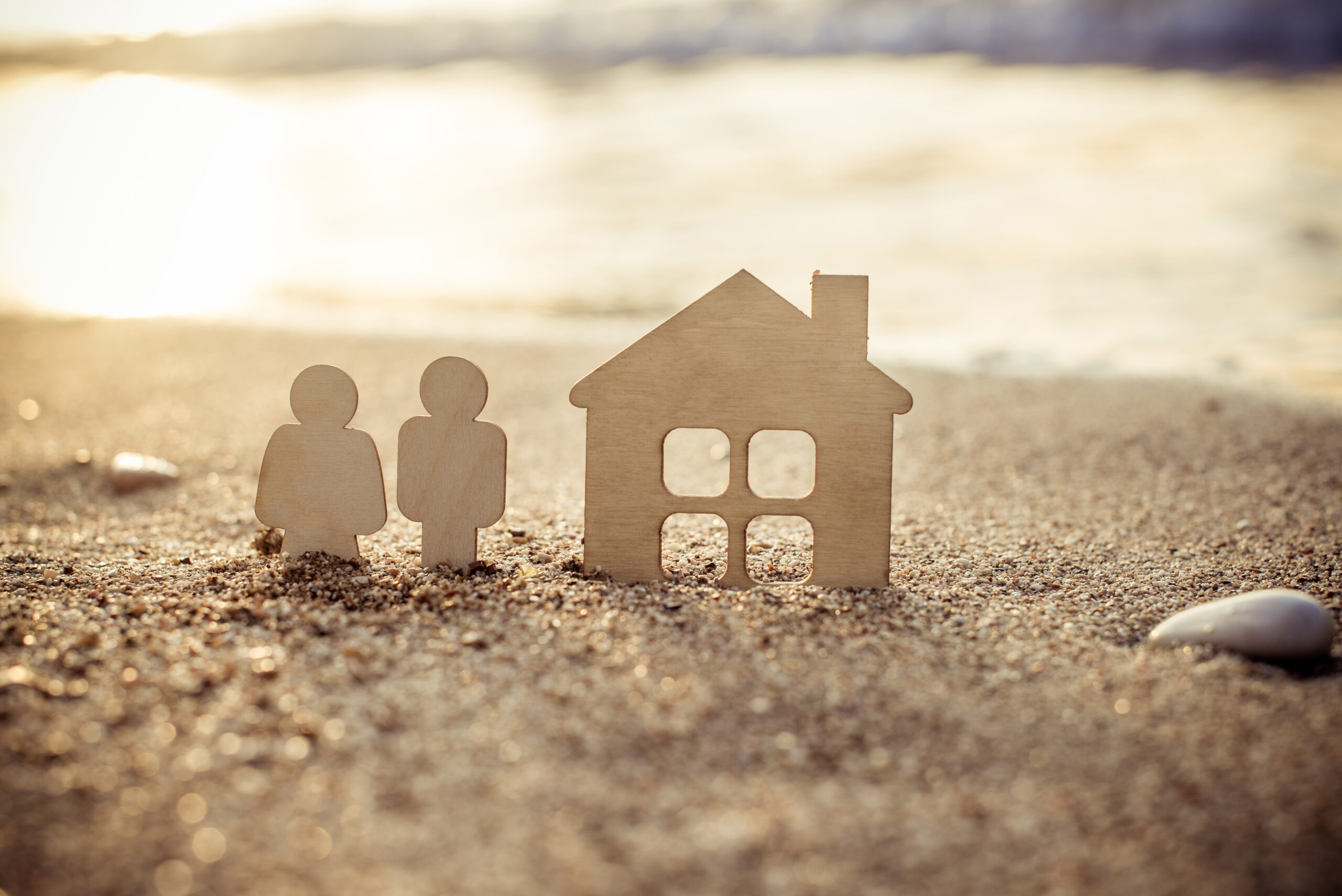 Holzfiguren von zwei Personen und Haus am Strand im Sonnenlicht.