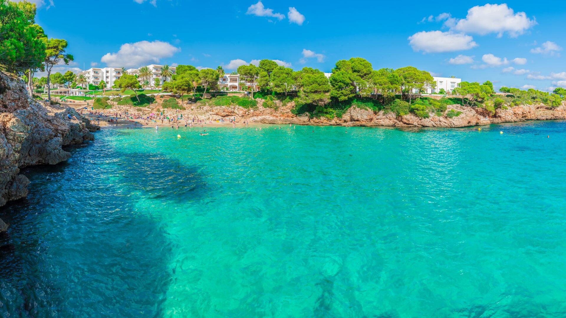 Türkises Wasser, Felsen, Bäume und weiße Häuser unter blauem Himmel.