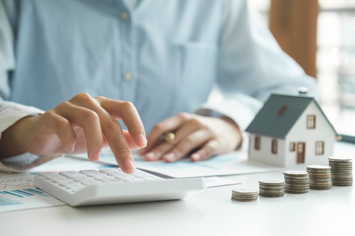 Mesch sitzt an Schreibtisch mit Tacshenrechner, Modellhaus und Münzen