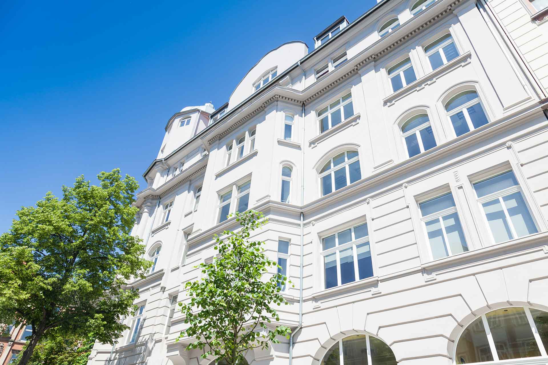 Weißes mehrstöckiges Wohnhaus mit Bogenfenstern und dekorativen architektonischen Details, gesehen an einem sonnigen Tag mit klarem blauen Himmel und grünen Bäumen im Vordergrund.
