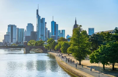 Moderne Wolkenkratzer und historische Gebäude säumen das Flussufer in einer Stadtansicht mit Menschen, die auf einem sonnigen Uferweg neben Bäumen spazieren gehen.