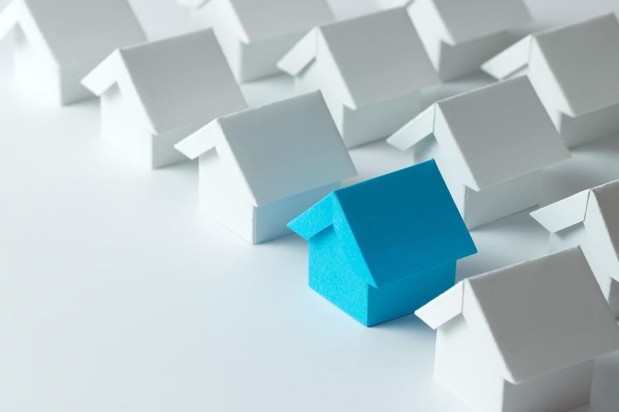 Eine Reihe kleiner weißer Papierhäuschen mit einem blauen Papierhäuschen in der Mitte, das symbolisiert, dass man auffällt oder anders ist.
