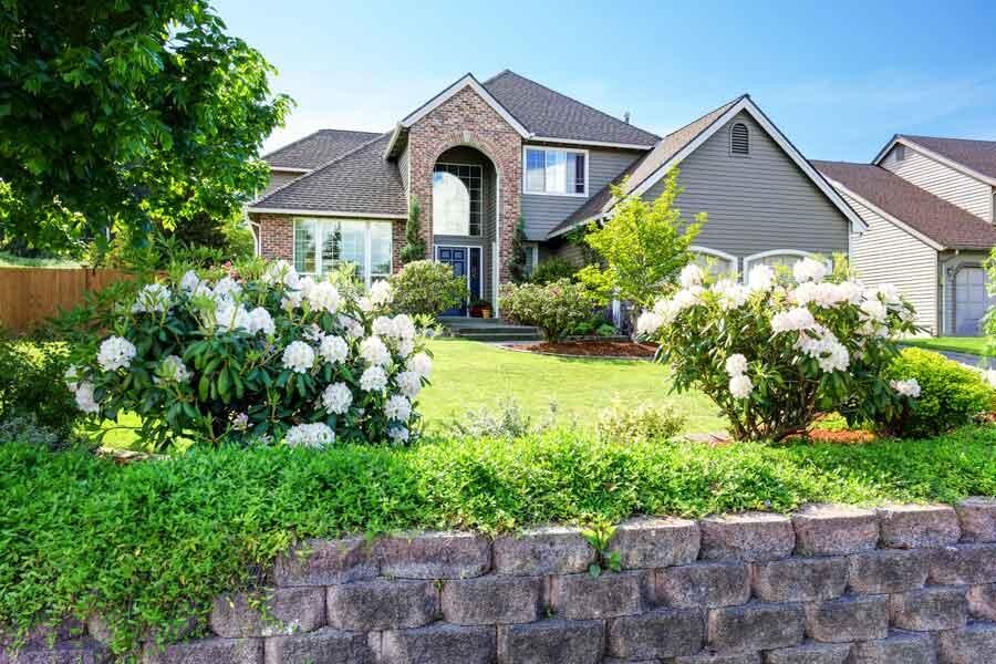 Zweistöckiges Vorstadthaus mit Ziegeln und Abstellgleisen, umgeben von einem gepflegten Rasen, blühenden Sträuchern und einer niedrigen Steinmauer an der Vorderseite.