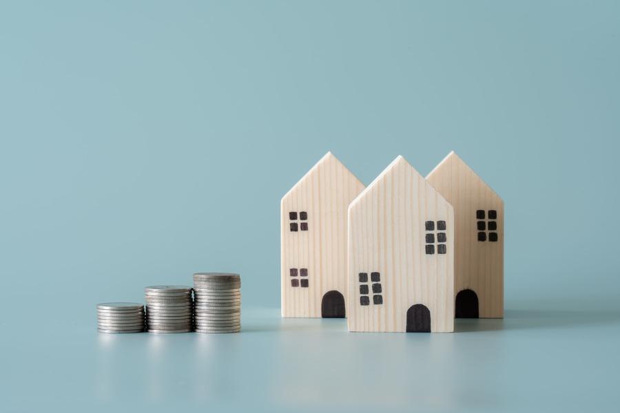 Drei kleine Holzhausmodelle stehen neben vier Stapeln von Münzen auf hellblauem Hintergrund.
