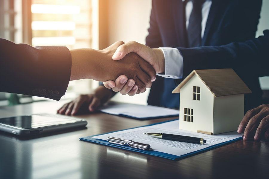 Zwei Personen in Geschäftskleidung schütteln sich die Hände über einem Schreibtisch mit einem kleinen Hausmodell, Dokumenten, einem Stift und einem Laptop, was auf einen Immobilien- oder Grundstücksvertrag schließen lässt.