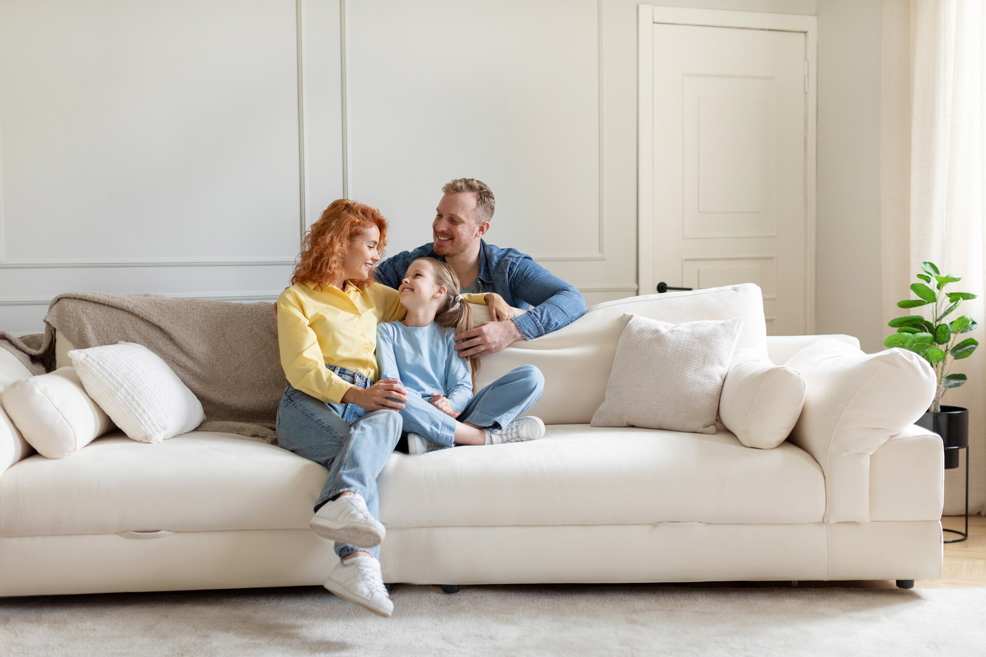Familie sitzt gemeinsam auf weißem Sofa im hellen Wohnzimmer.