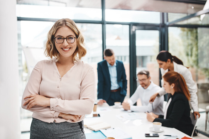Frau mit Brille steht vor Kollegen am Besprechungstisch