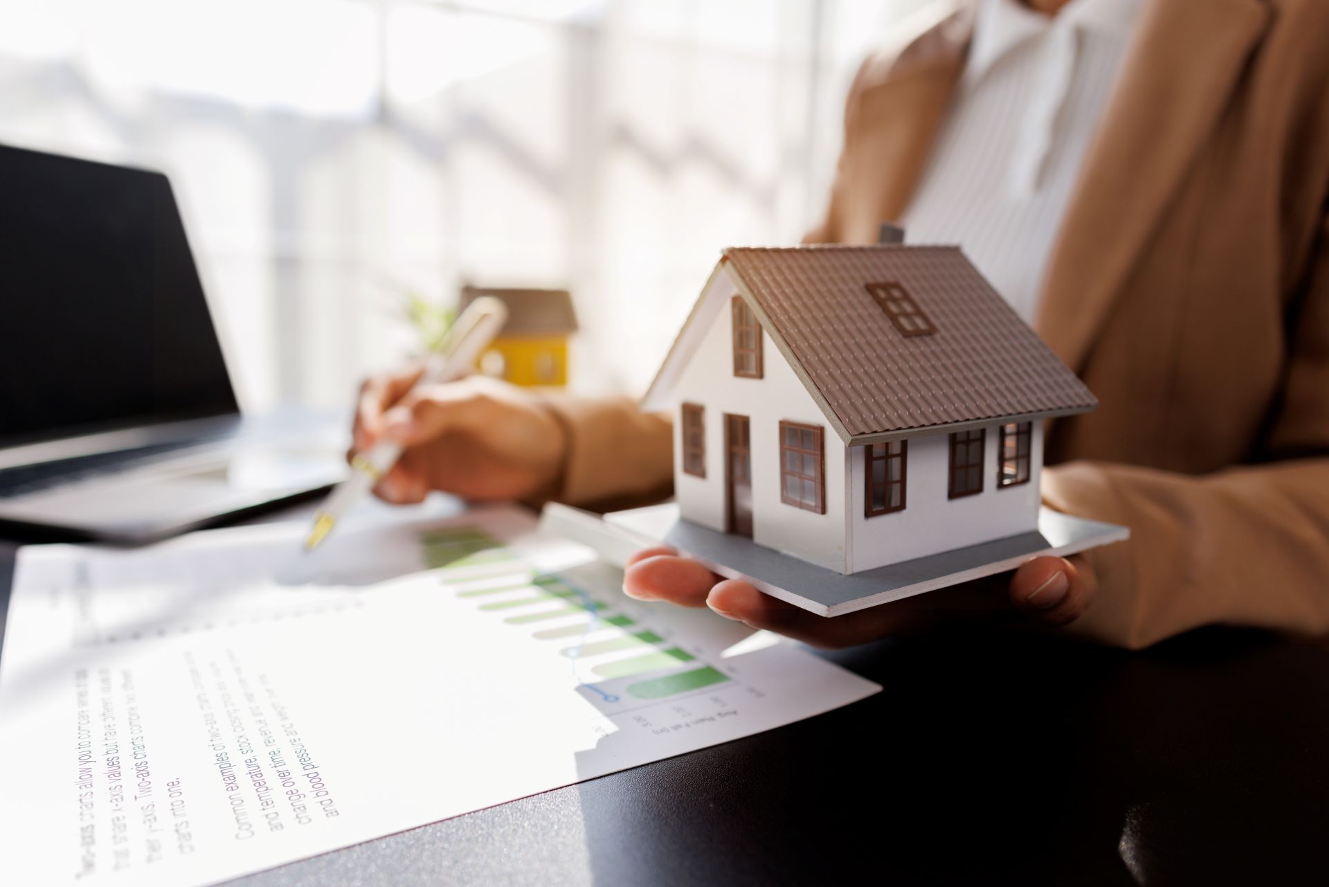 Person mit Modellhaus, Dokumenten und Laptop am Schreibtisch.