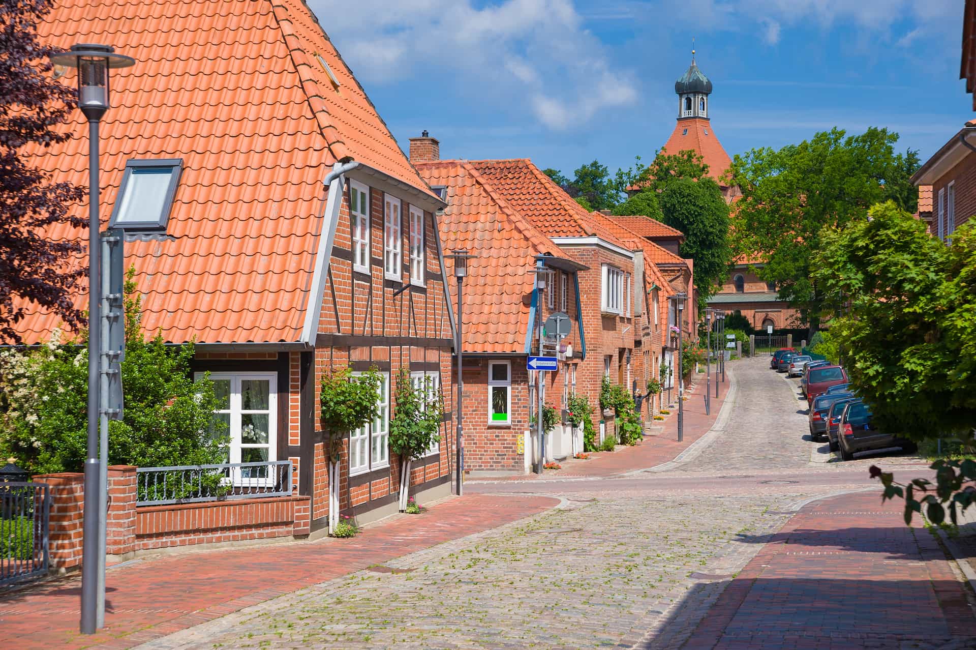 Kopfsteinpflasterstraße mit Backsteinhäusern, roten Dächern und geparkten Autos.