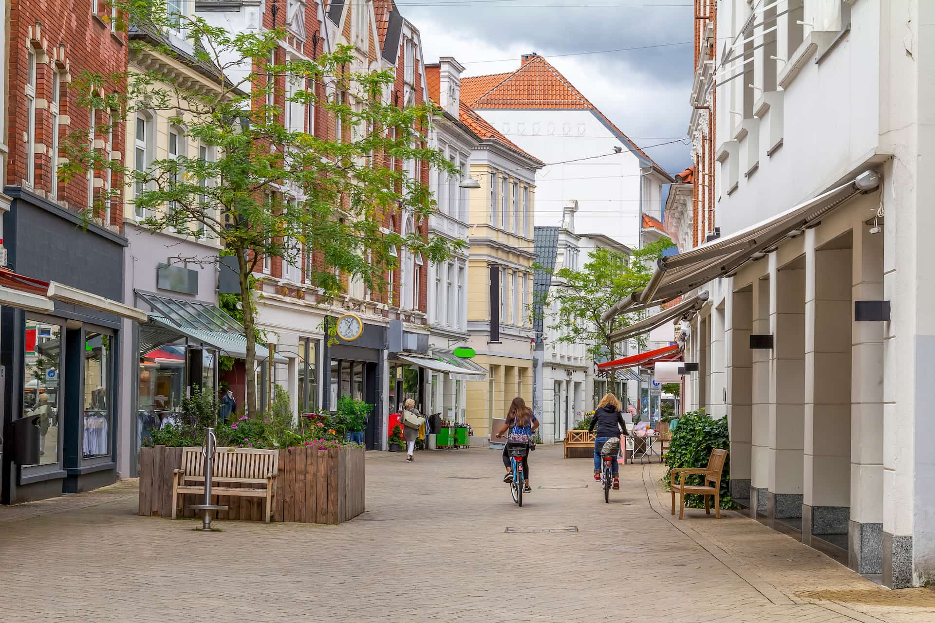 Radfahrer auf ruhiger Straße mit Bäumen, Läden und Cafés.