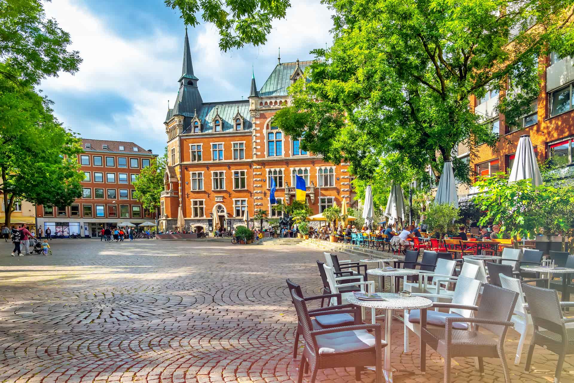 Platz mit Café-Sitzplätzen, Bäumen und historischem Gebäude.