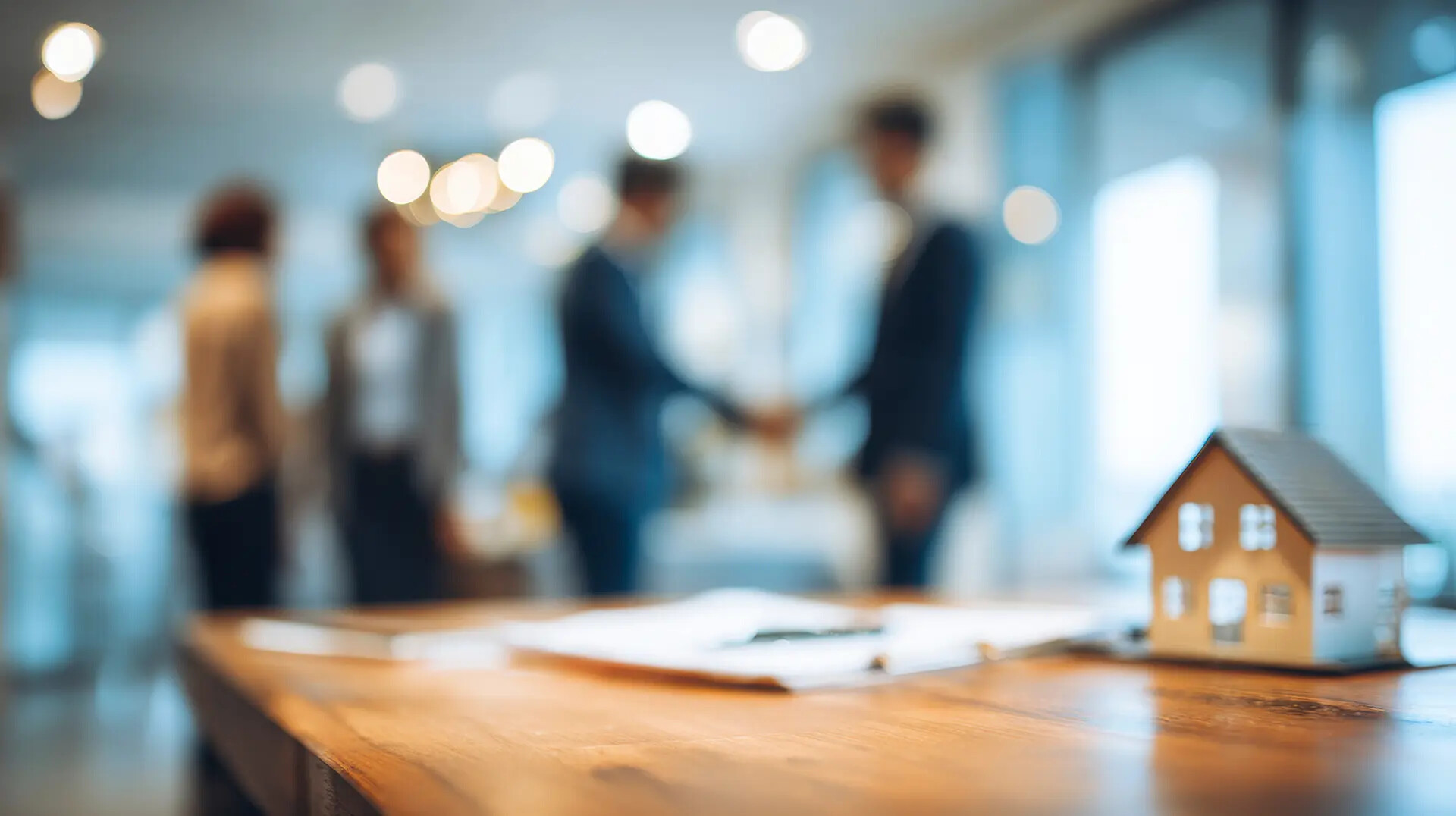 Innenaufnahme mit einem Immobilienvertrag, verschwommenen Figuren, die sich die Hand geben, und einem Miniaturhaus auf einem Holztisch. Verschwommener Hintergrund, geschäftliches Umfeld