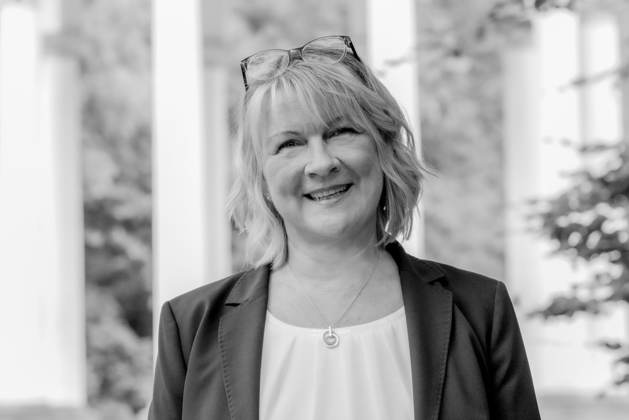 Frau mit Brille auf dem Kopf, Blazer, im Freien bei verschwommenen Säulen.