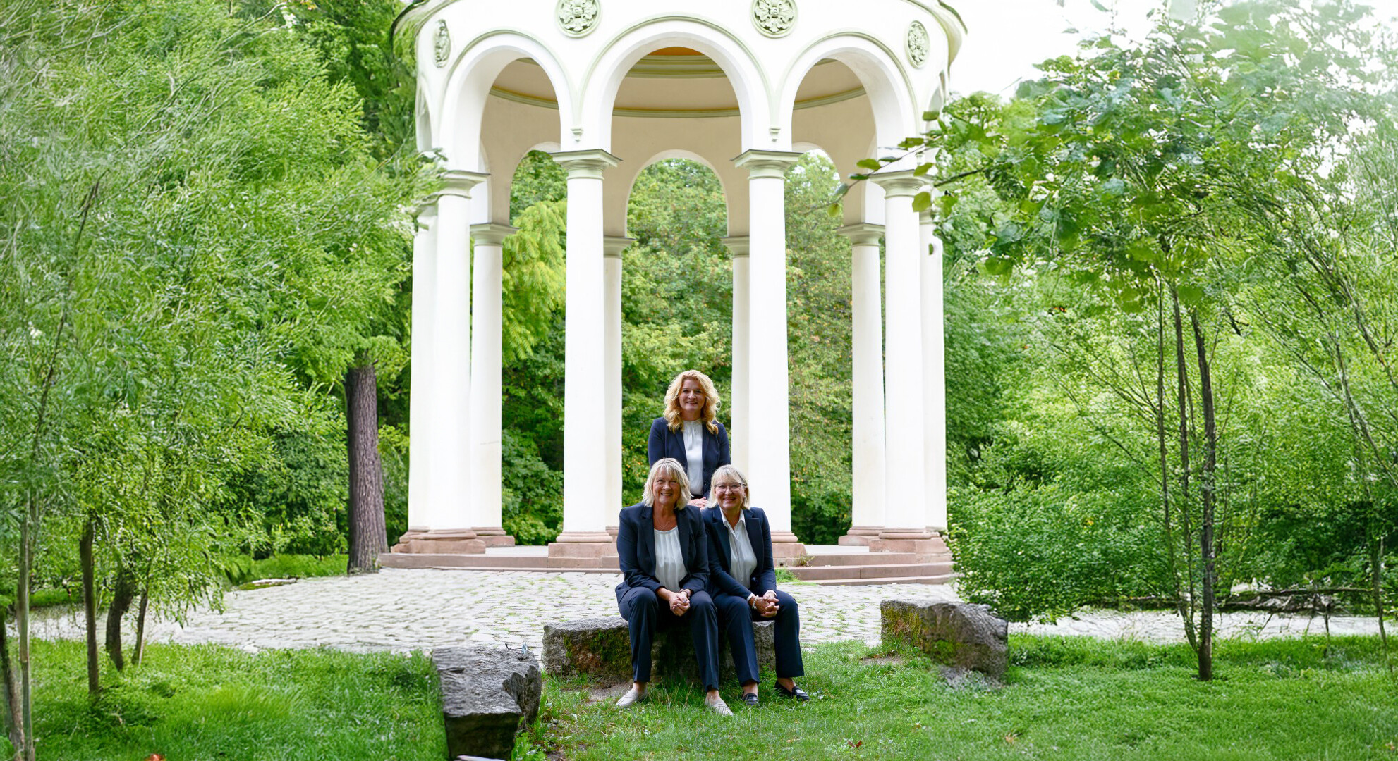 Drei Frauen in Businesskleidung vor weißem Pavillon im Grünen.