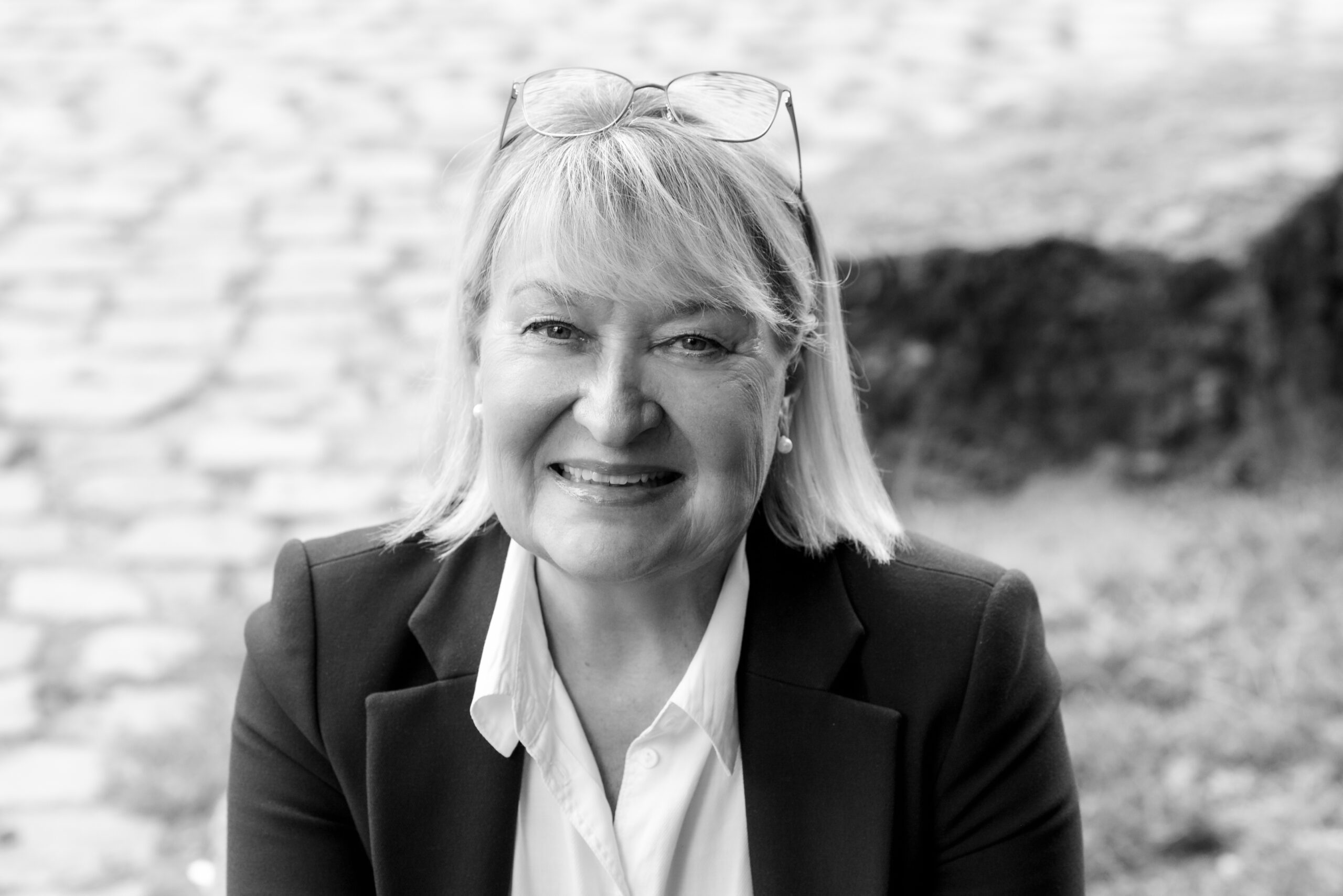 Frau mit Brille im Blazer sitzt draußen auf Steinweg.