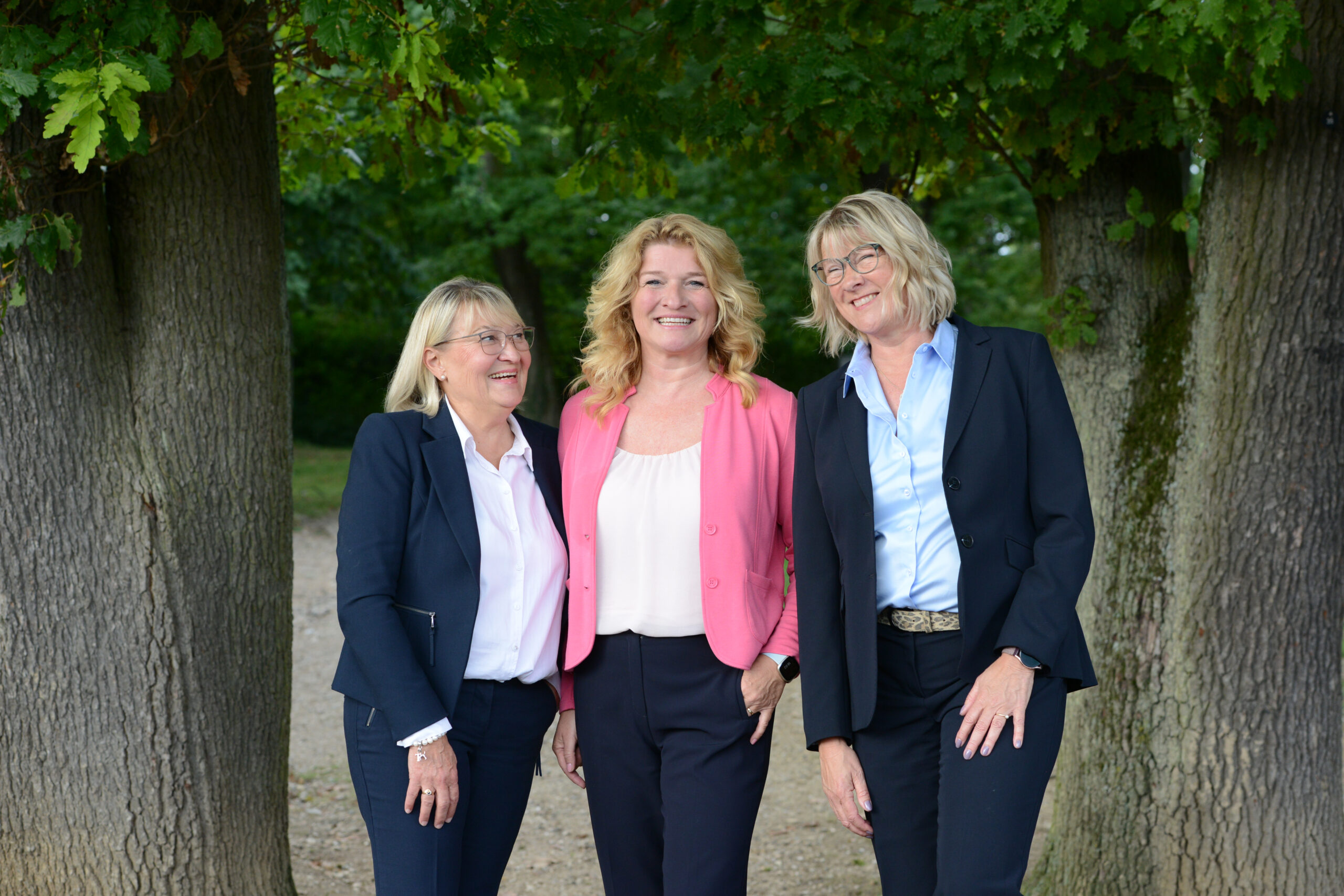 Drei geschäftlich gekleidete Frauen lächeln draußen zwischen Bäumen.