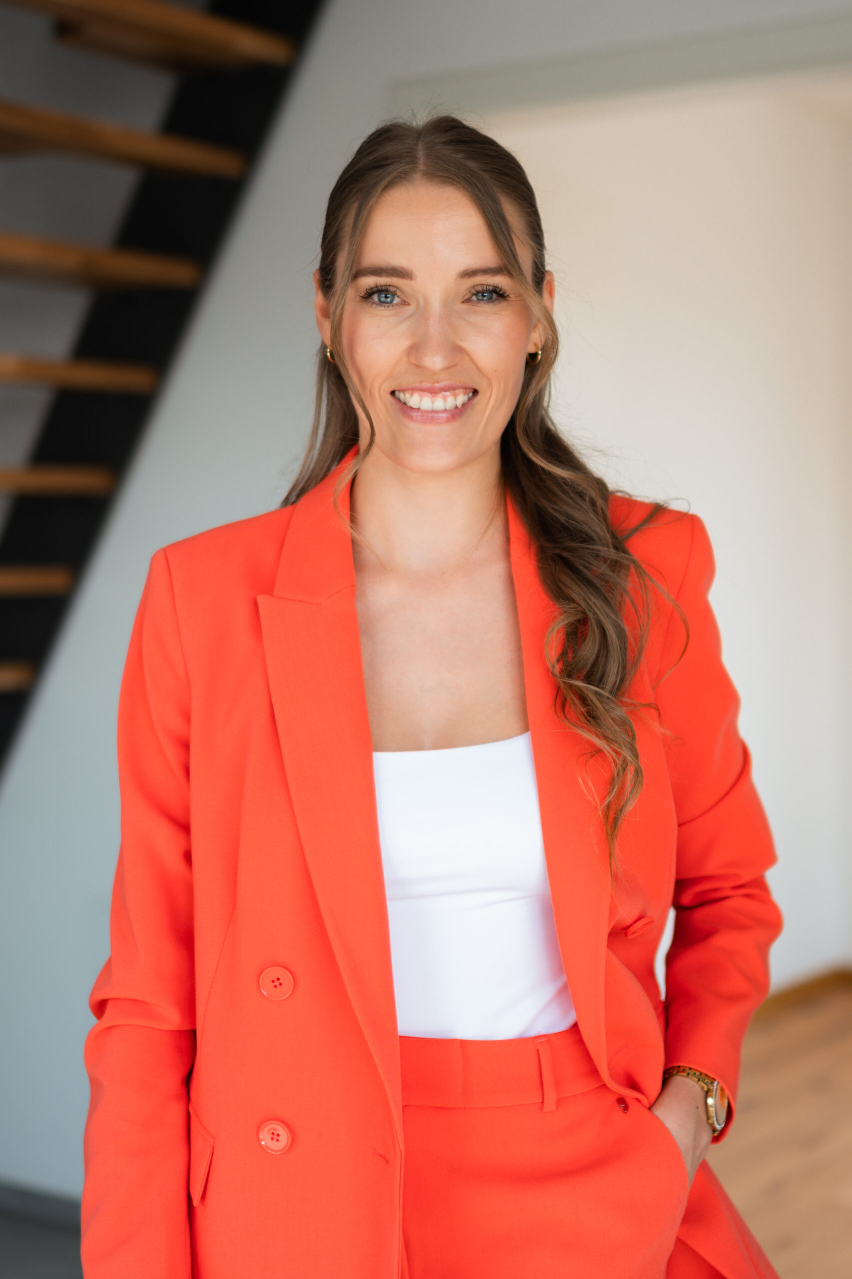 Frau lächelnd im orangefarbenen Anzug steht drinnen bei einer Treppe. Sie ist für Marketing und Kommunikation bei Doldi Immobilie zuständig.