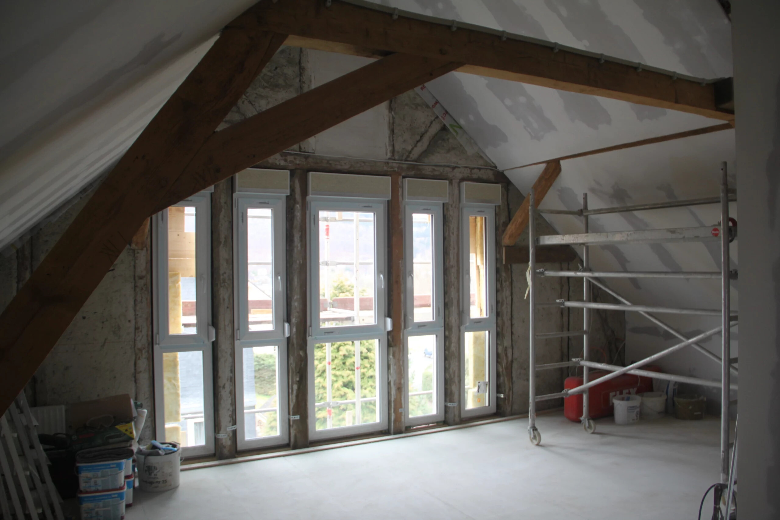 Unausgebauter Dachboden mit großen Fenstern, Balken und Gerüsten.