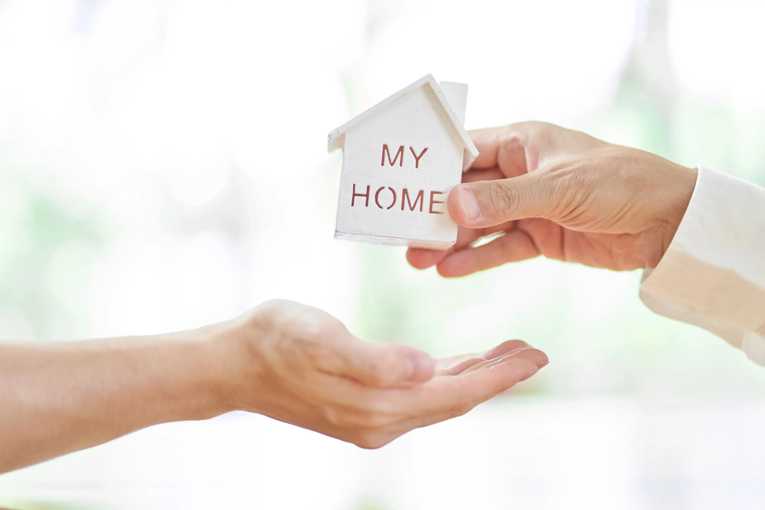 Hand überreicht Modellhaus mit "MY HOME" an offener Hand.