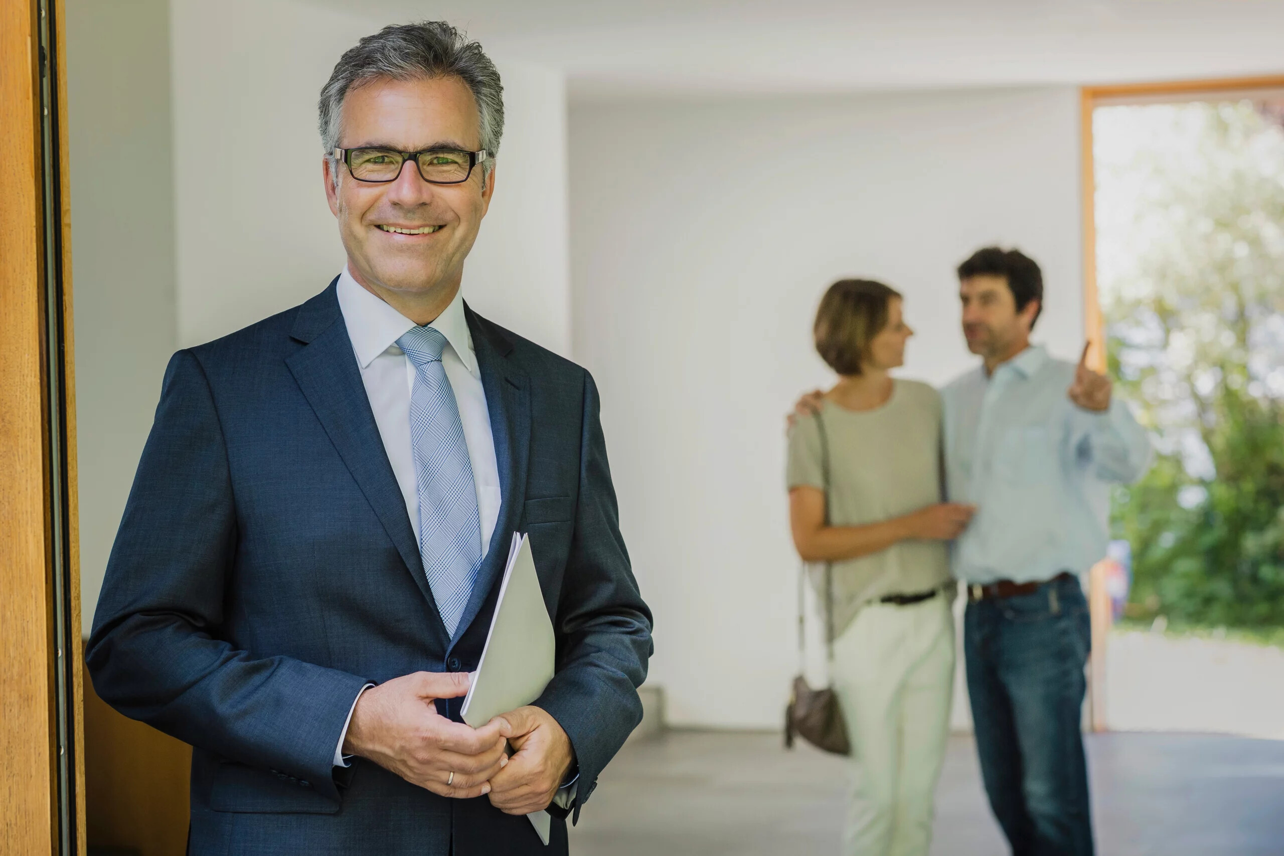 Mann im Anzug mit Ordner, Paar im Hintergrund eines hellen Raums.