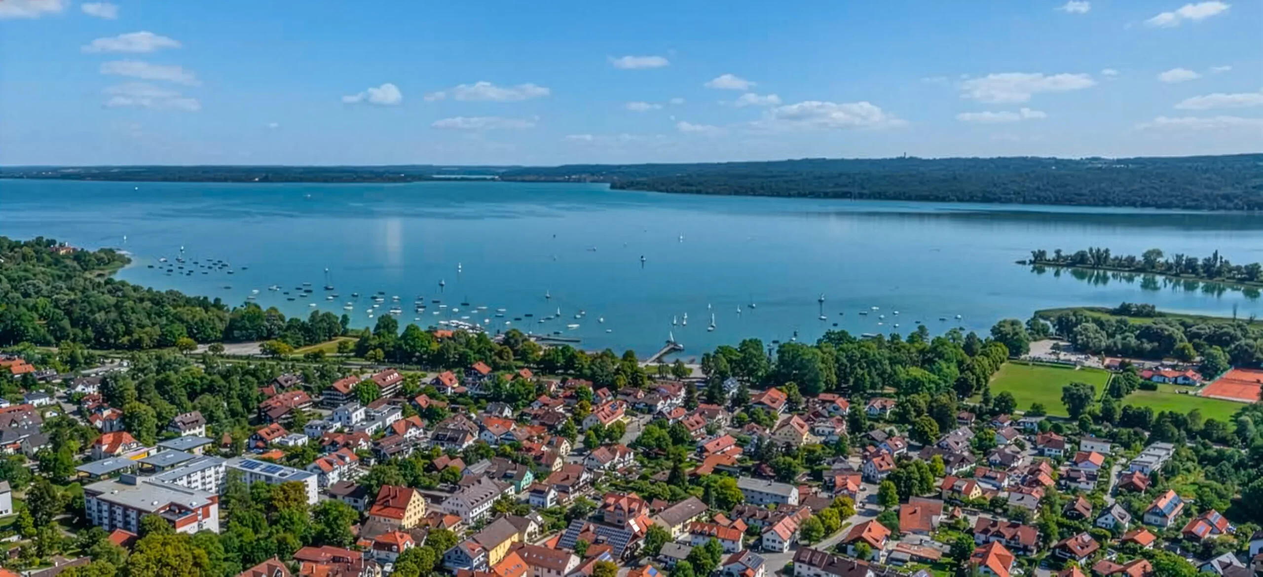 Stadt am See, rote Dächer, Boote, bewaldete Hügel, blauer Himmel.