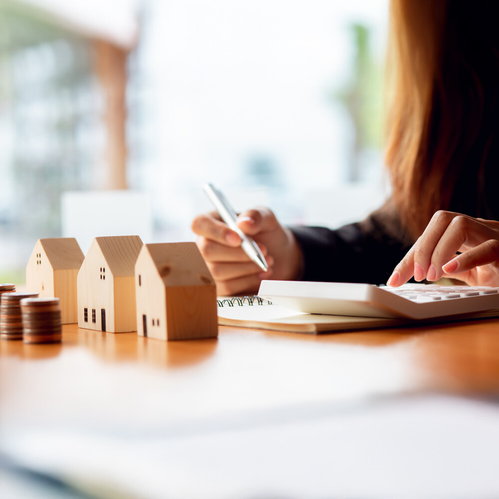 Hände rechnen am Taschenrechner, lleine Holzhäuser und Münzenstapel