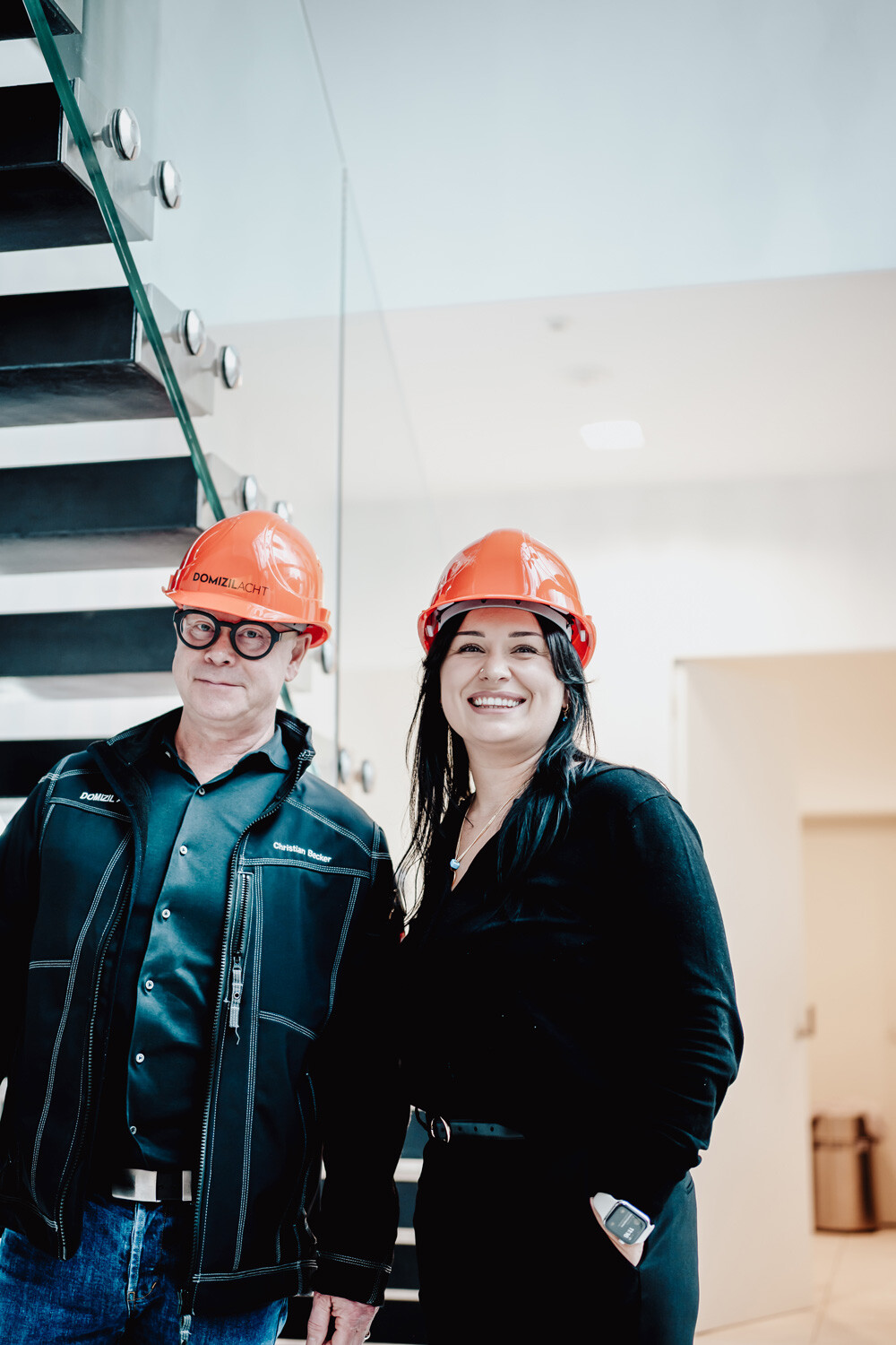 Zwei Personen mit orangefarbenen Helmen lächeln auf einer Treppe.