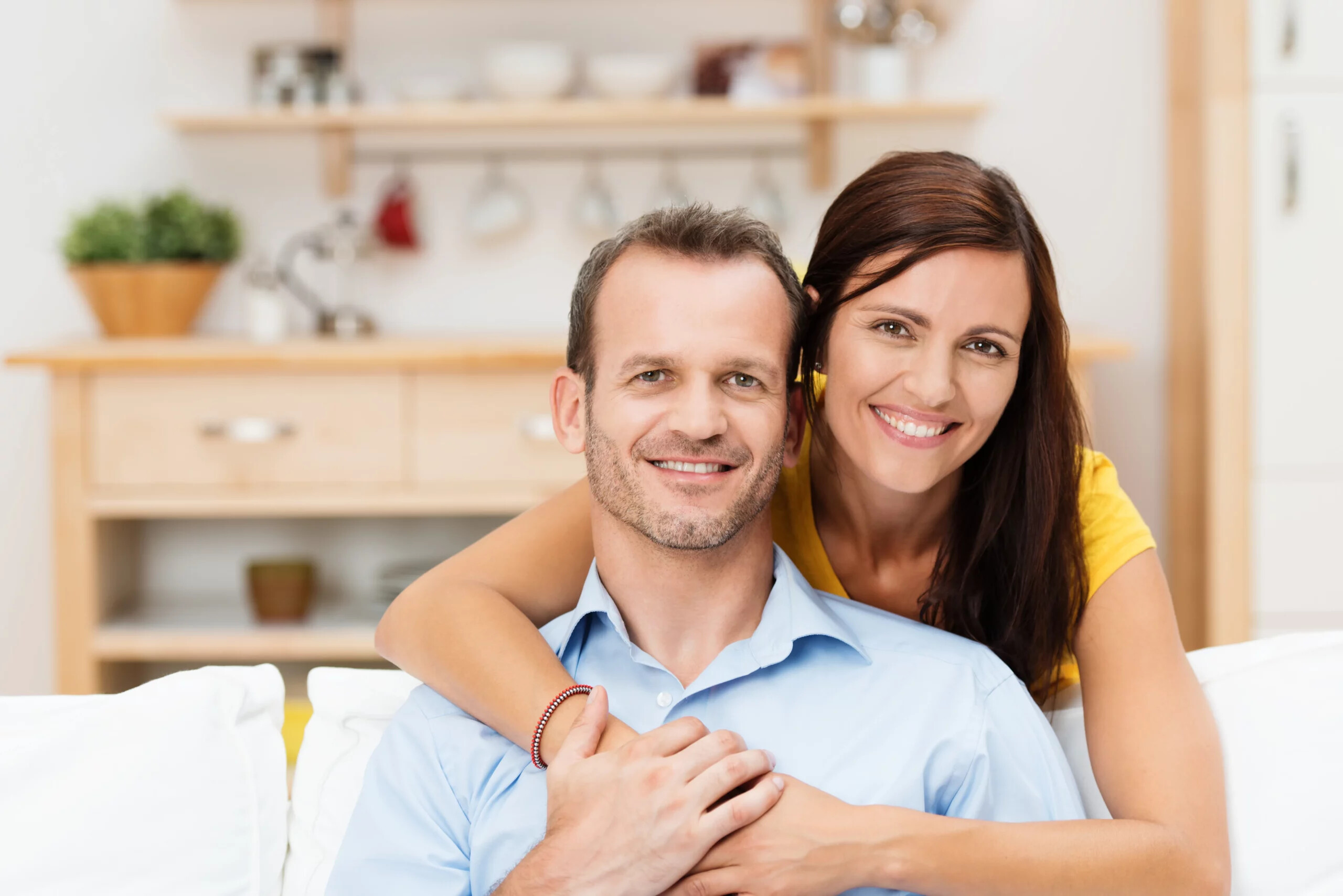 Frau umarmt Mann auf Sofa in heller, gemütlicher Küche.