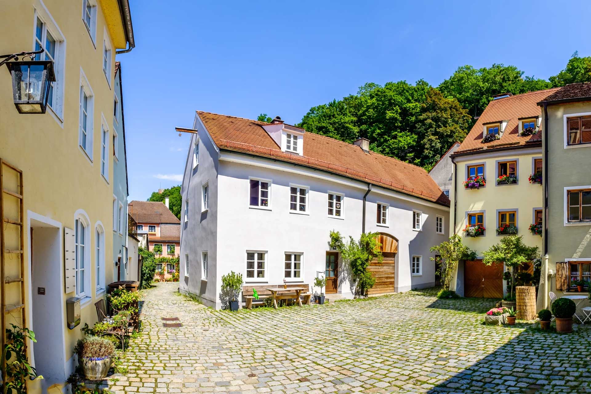 Bunter Hof mit Kopfsteinpflaster, roten Dächern, Topfpflanzen.