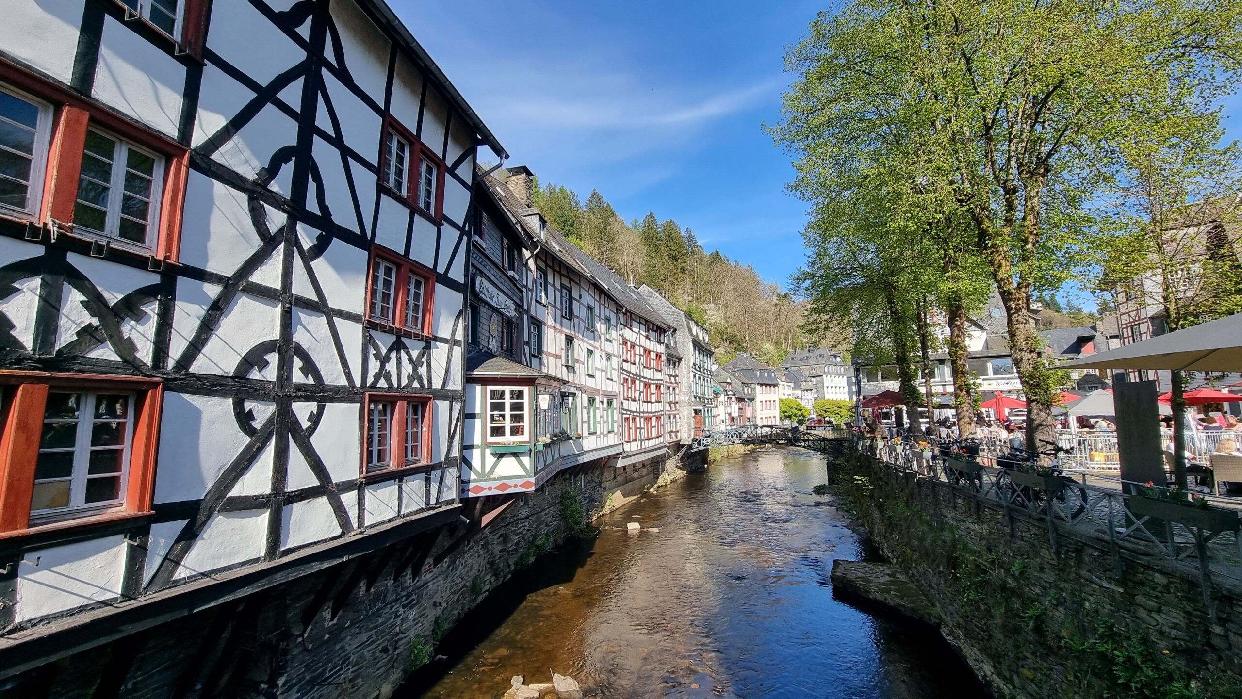 Fachwerkhäuser am Fluss mit Bäumen und Cafés bei blauem Himmel.