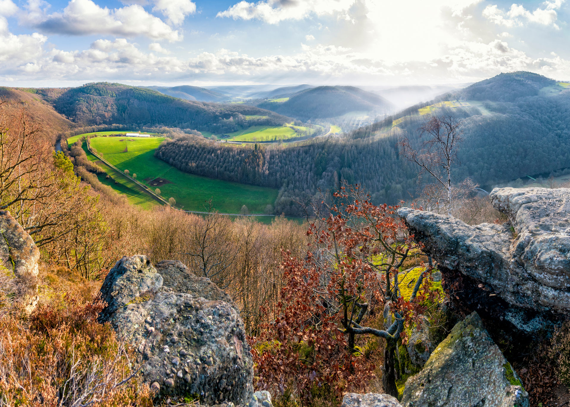 Sanfte Hügel, Wald und Fluss unter sonnenbeschienenem Himmel.