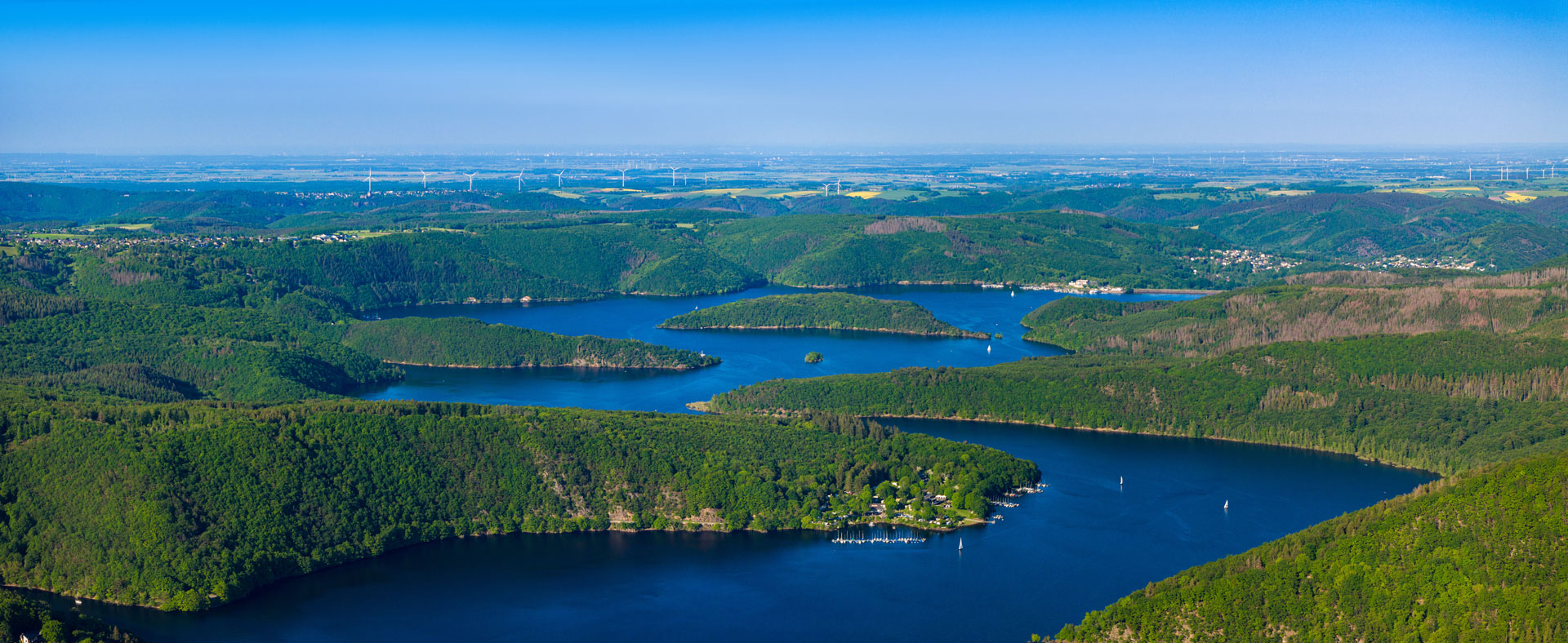 Luftaufnahme eines sich windenden blauen Sees inmitten grüner Hügel unter klarem Himmel.