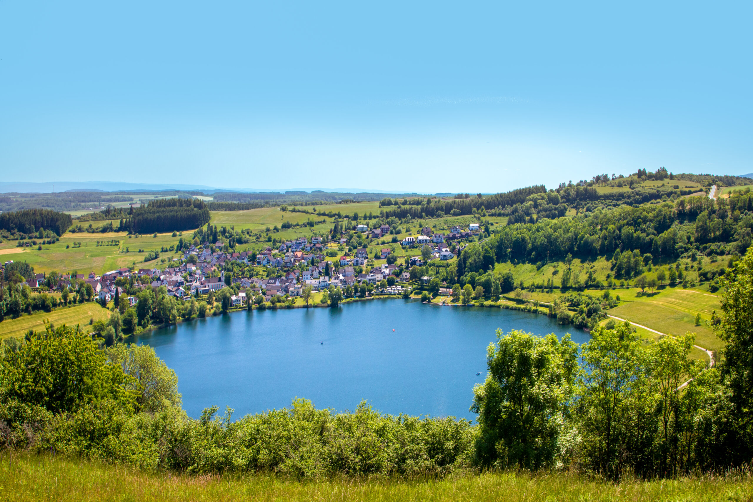 Kleinstadt und Wälder umgeben einen runden See im Sonnenschein.