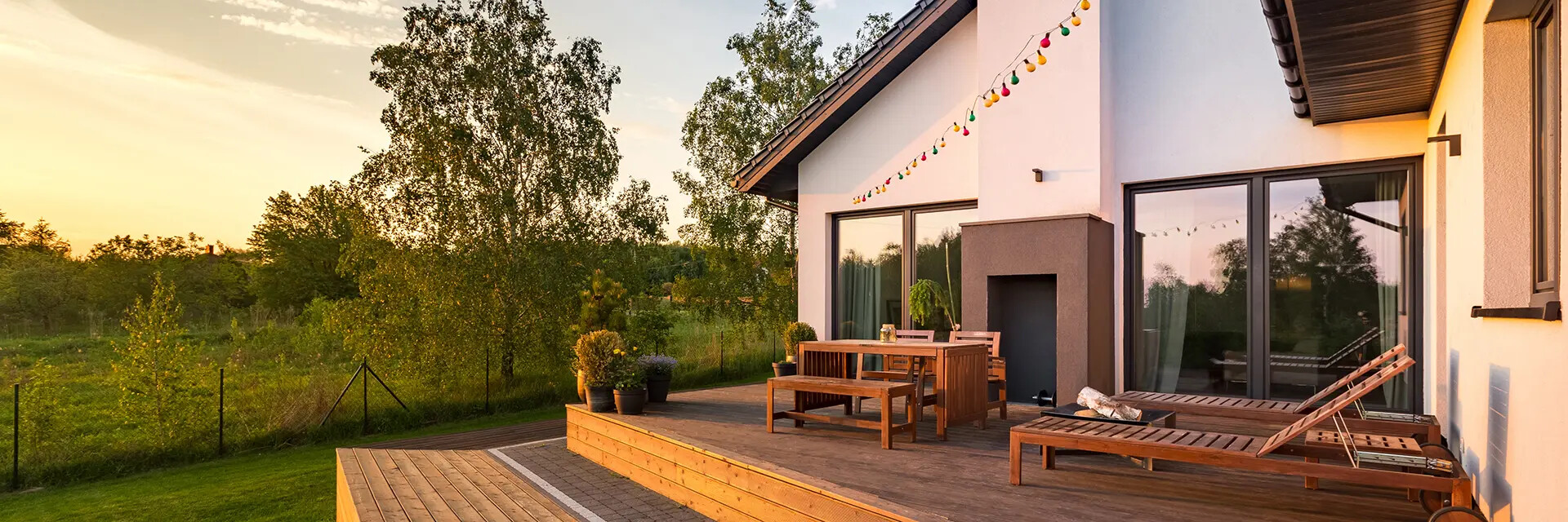 Patio mit Holzmöbeln, Lichterkette und Blick auf Bäume bei Sonnenuntergang.