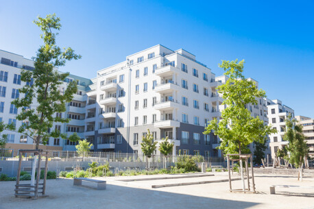 Weißes Wohngebäude mit Balkonen, junge Bäume, blauer Himmel.