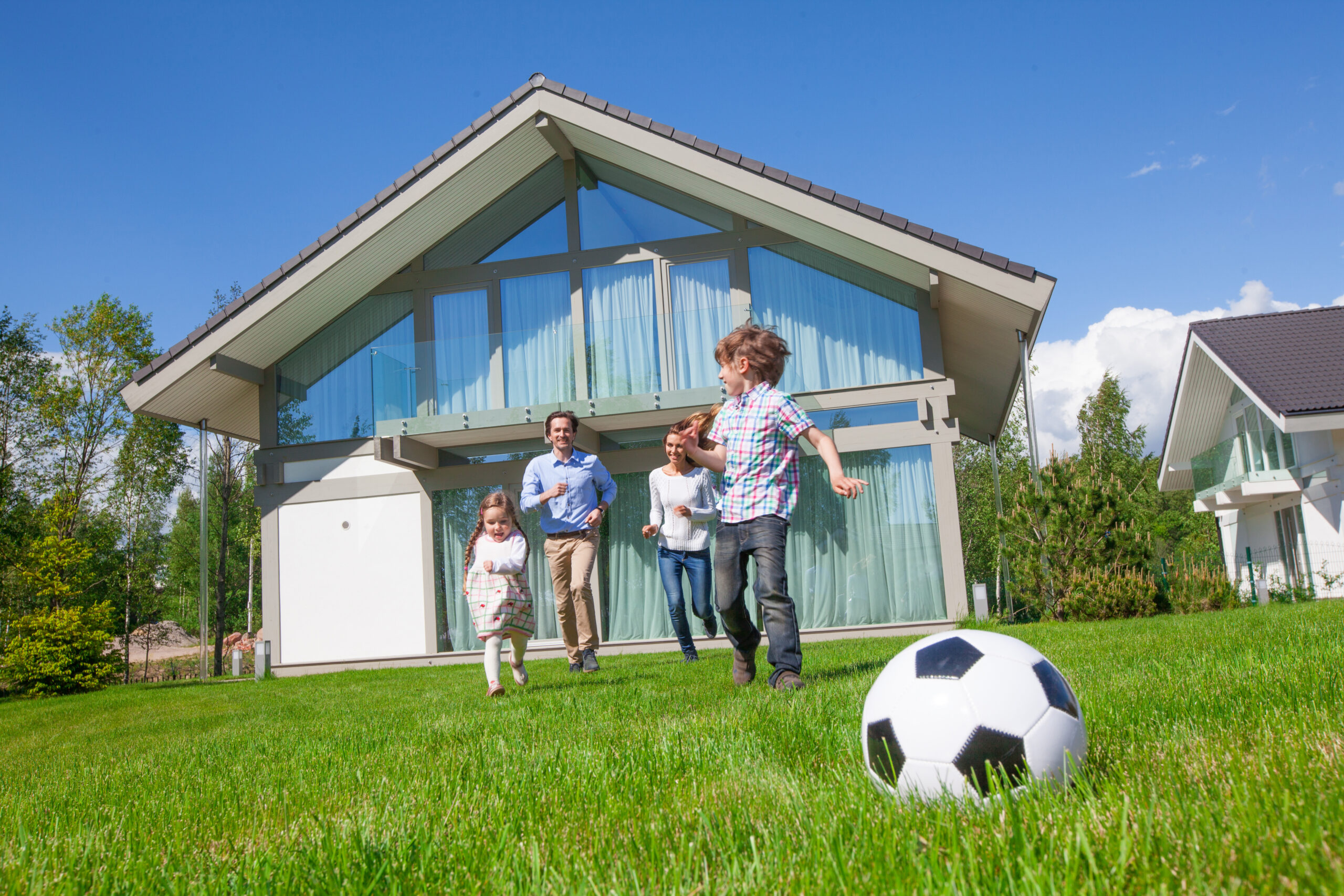 Vierköpfige Familie läuft auf Rasen, modernes Haus, Fußball vorn.