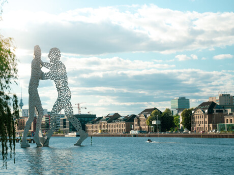Zwei Figuren als Skulptur im Fluss, Stadt und Boot dahinter.