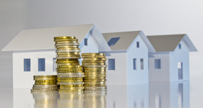 Münzstapel vor drei kleinen Modellhäusern auf spiegelnder Fläche.