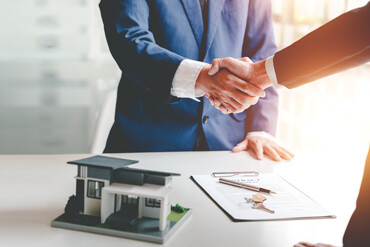 Geschäftsleute geben sich vor Vertrag, Schlüsseln und Minihaus die Hand.