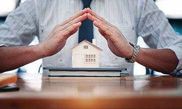 Die Hände bedecken ein kleines Modellhaus auf einem Tisch, das Schutz bietet.