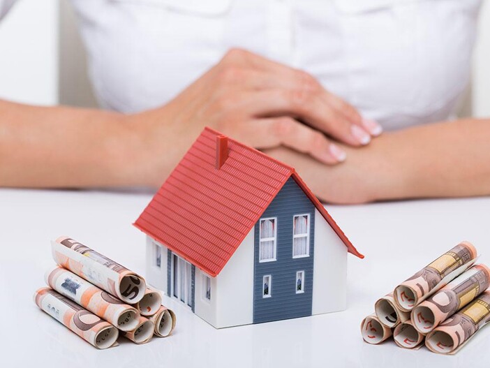 Modellhaus mit Euro-Scheinen und gefalteten Händen auf Tisch.