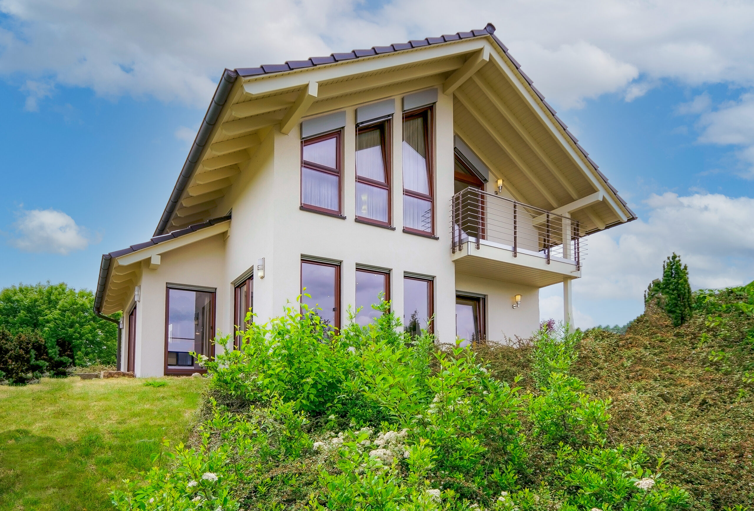 Zweistöckiges Haus mit großen Fenstern, Balkon und viel Grün.
