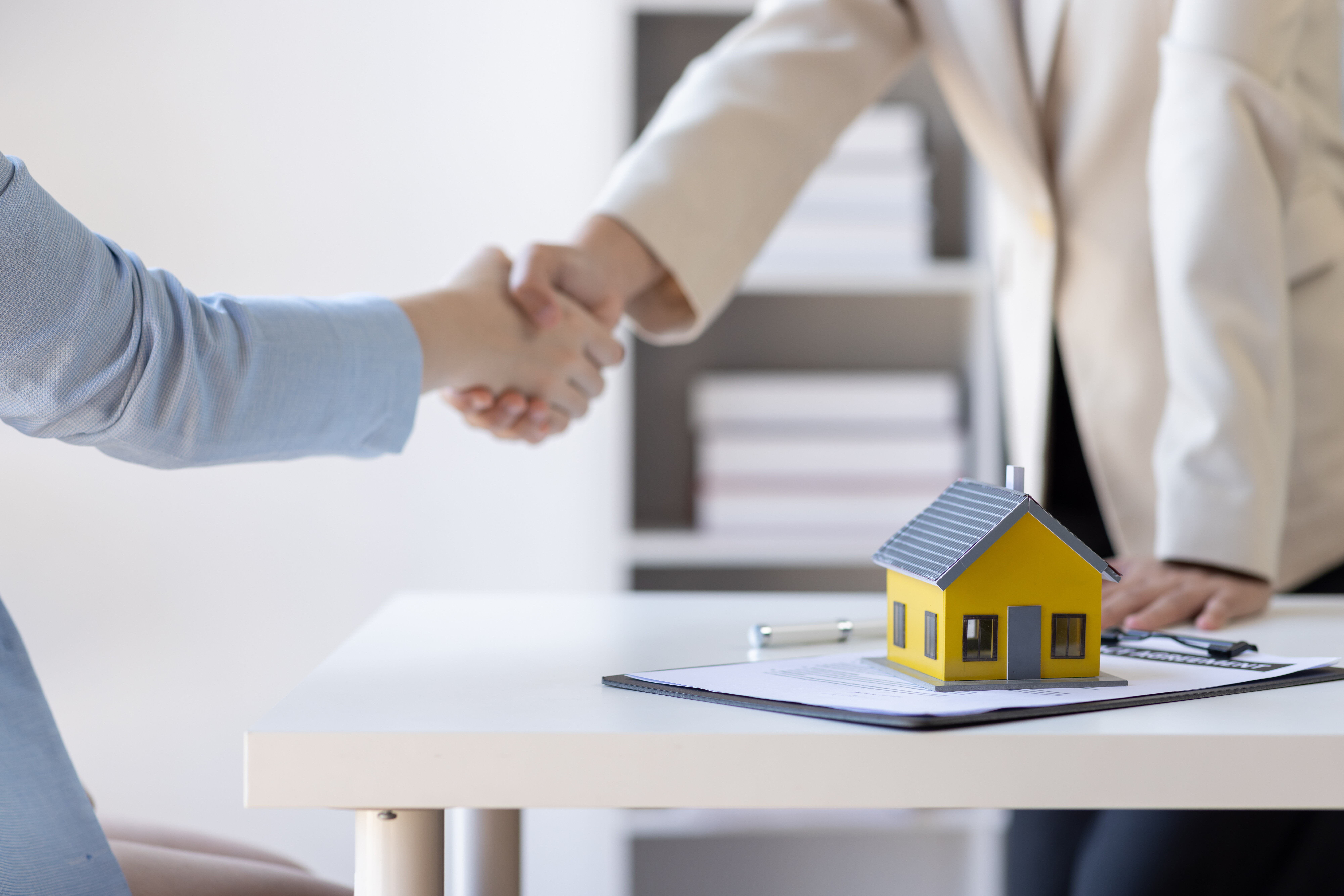 Händedruck über Schreibtisch mit Miniaturhaus und Unterlagen.