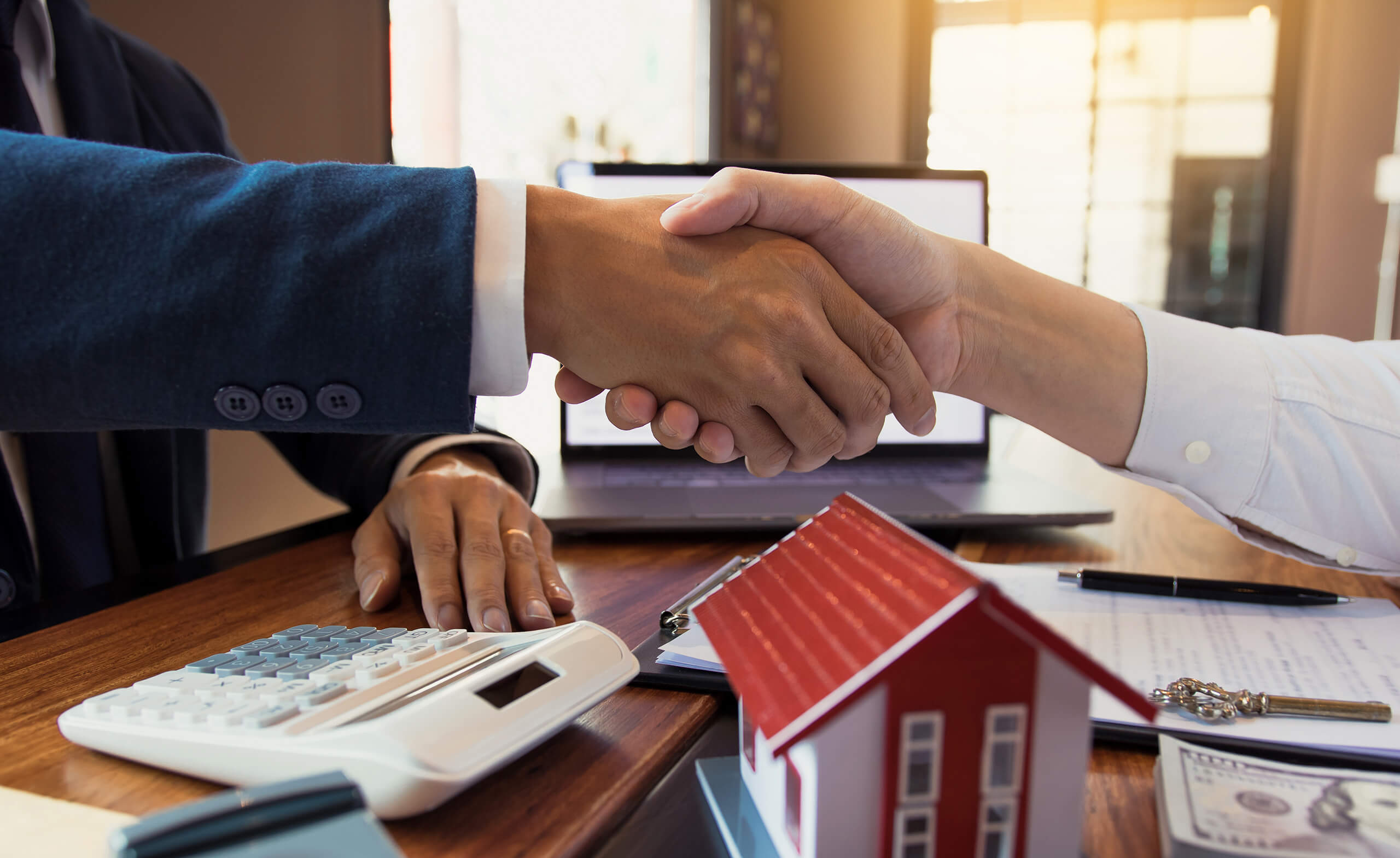 Händedruck über Schreibtisch mit Hausmodell, Rechner und Dokumenten: Symbol für Verkauf mit Makler