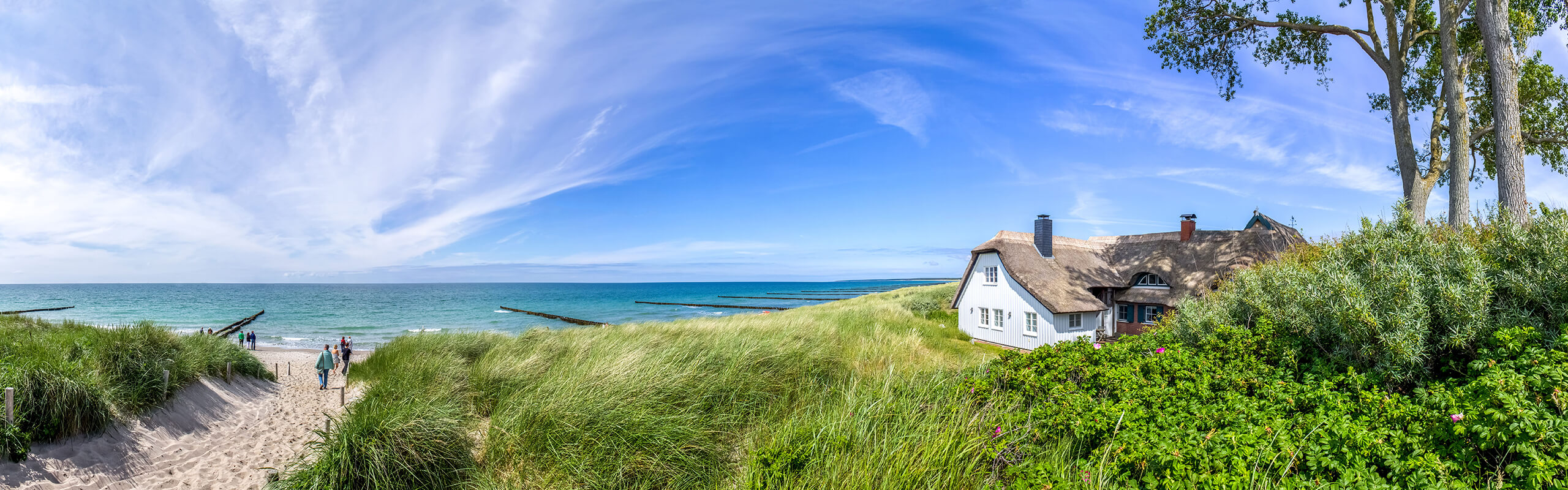 Reetdachhaus, grasbewachsene Dünen, Menschen auf dem Weg zu Strand und Meer.