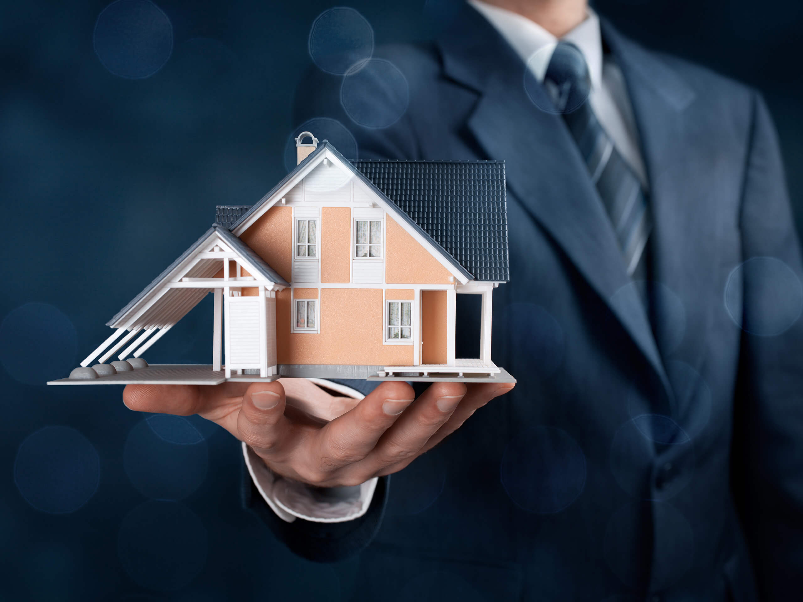 Mann im Anzug hält Modellhaus vor blauem Hintergrund.